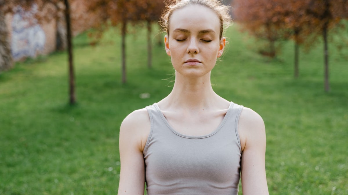 Young woman meditating outdoors in a lush green park during daytime, promoting mindfulness and relaxation.